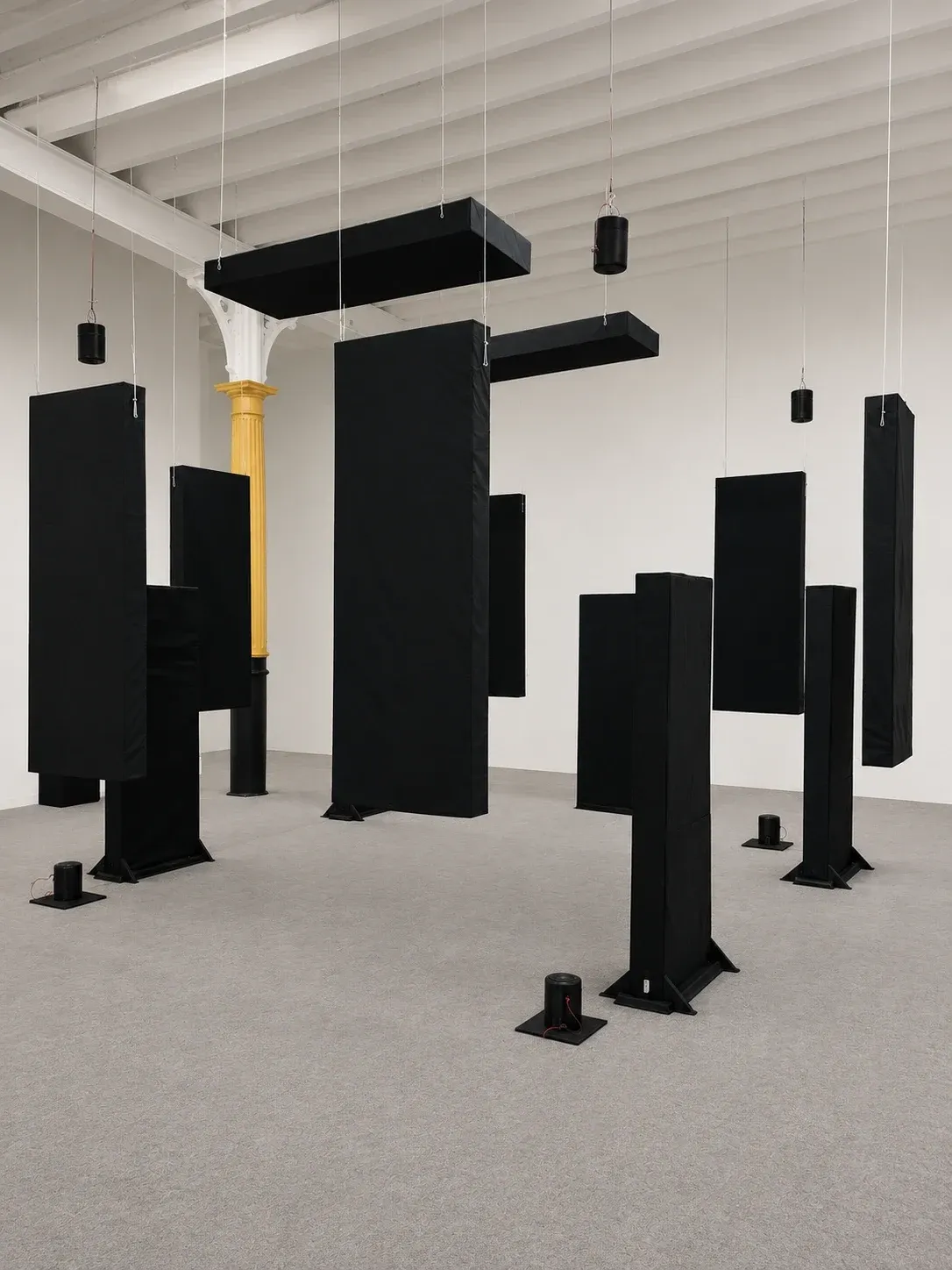 The Indifference installation, black fabric-covered panels hanging from the ceiling and standing on the floor of a white gallery, with small black speakers between them.