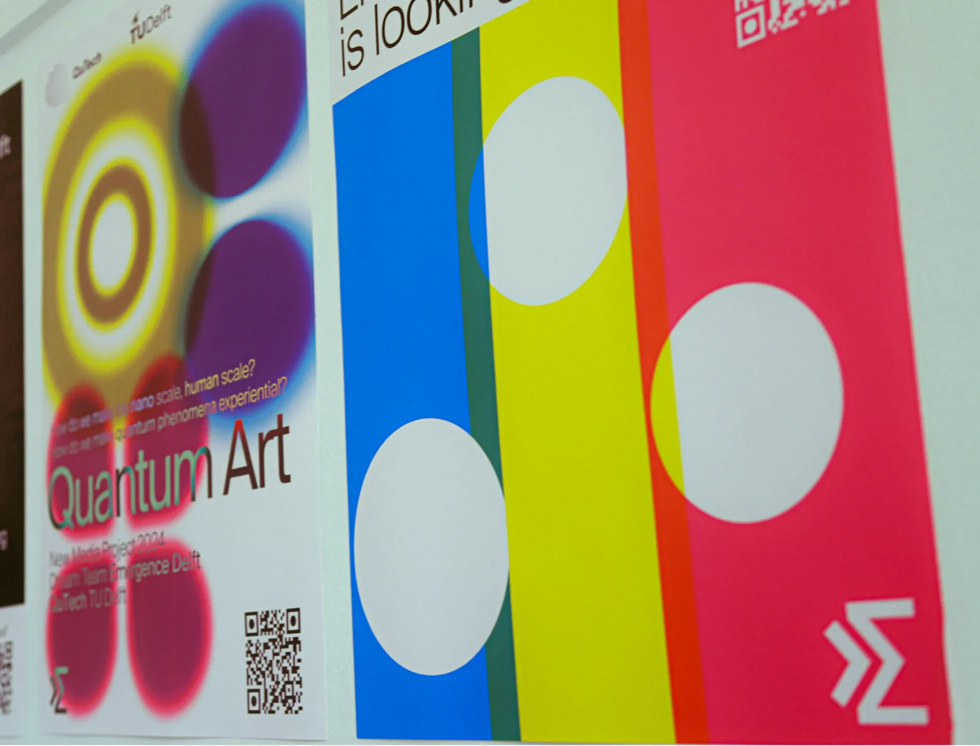 Three Emergence Delft posters on a wall, one with a dark spiral, one with glowing yellow and purple circles titled Quantum Art, and one with blue, yellow and pink stripes and white circles.