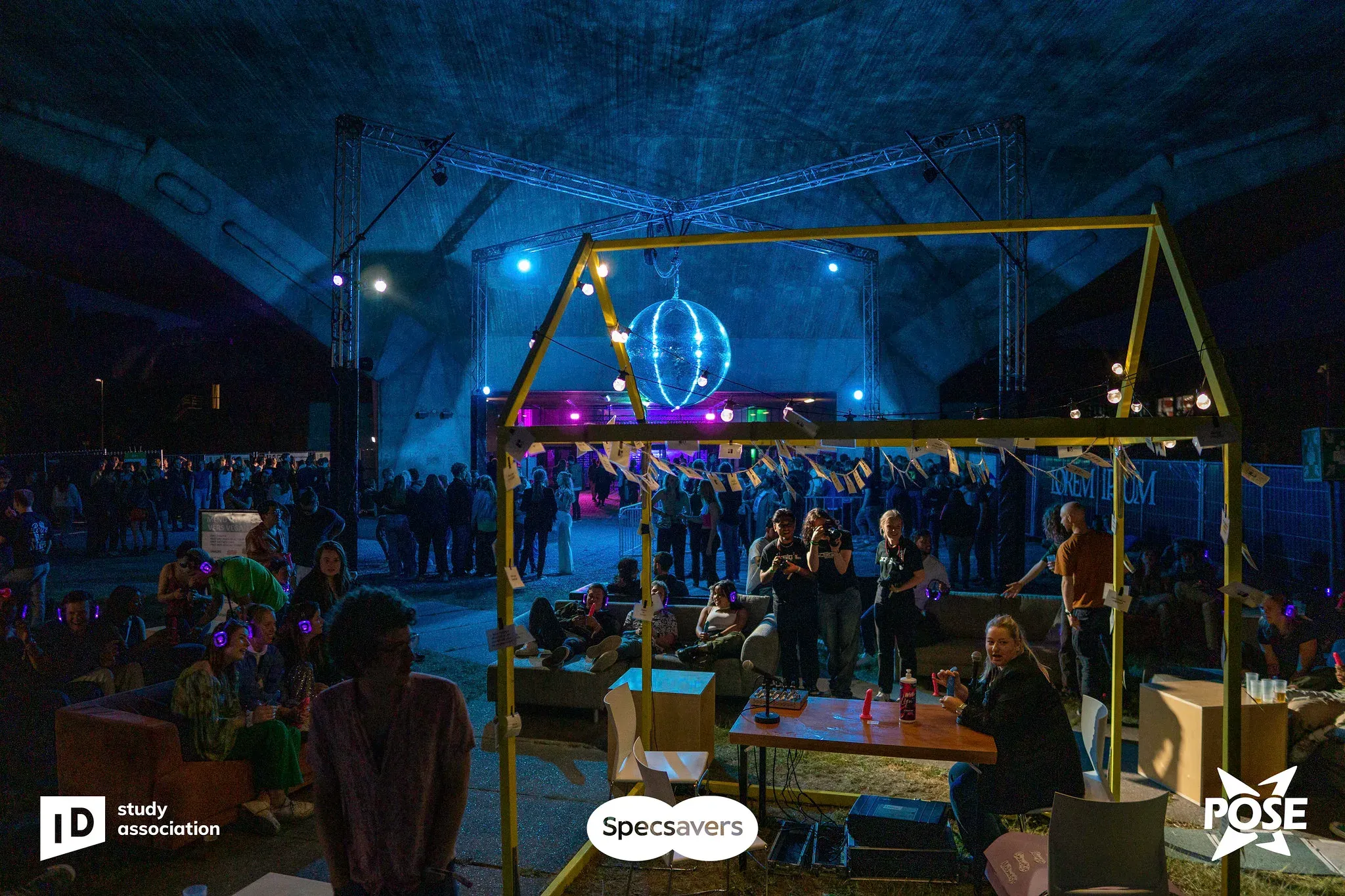 The festival at night, with a crowd around a stage framed by a yellow house-shaped structure and a mirror ball.
